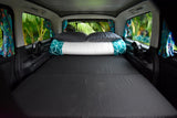 Spacious RV rental bed set up with pillows, fans, and tropical scenery visible through side windows. Ideal for nature escapes.