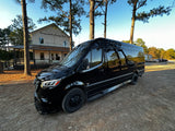 A sleek black Mercedes-Benz Sprinter rv rental van parked on a dirt lot near "Sasquatch's Candy Den." Tall pine trees frame the scene.