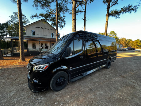 A sleek black Mercedes-Benz Sprinter rv rental van parked on a dirt lot near "Sasquatch's Candy Den." Tall pine trees frame the scene.