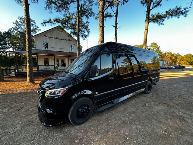 A sleek black Mercedes-Benz Sprinter rv rental van parked on a dirt lot near "Sasquatch's Candy Den." Tall pine trees frame the scene.