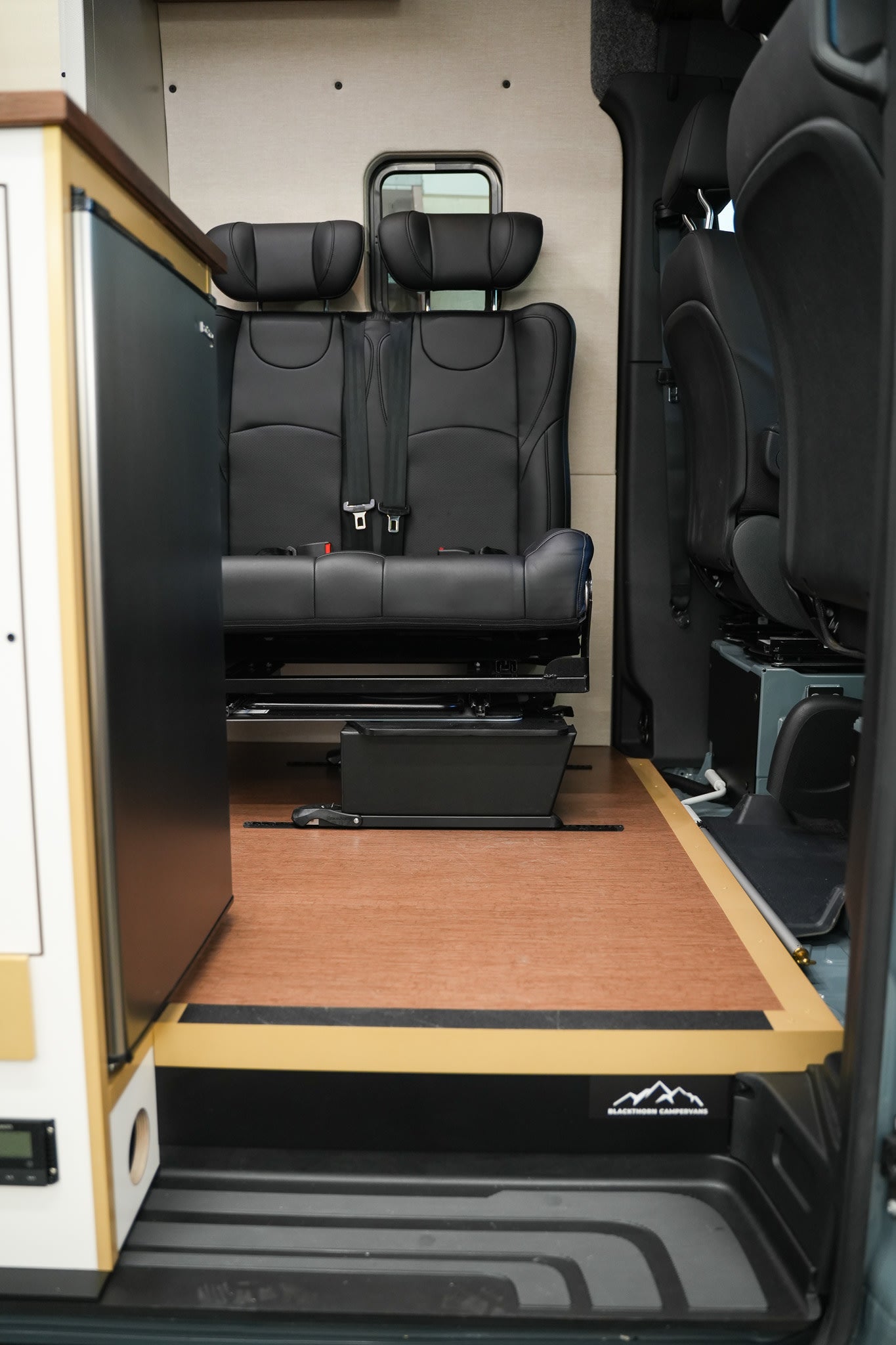 Inside an rv rental: comfortable black passenger seats, wooden floor, and a kitchen cabinet on the left.