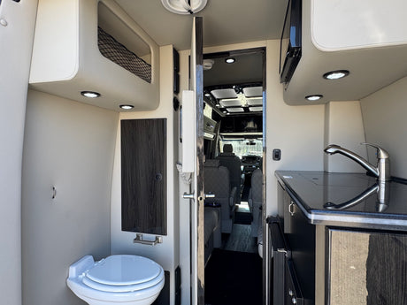 RV rental interior: compact kitchen (sink, microwave), toilet, storage nets, and door to cab seats.