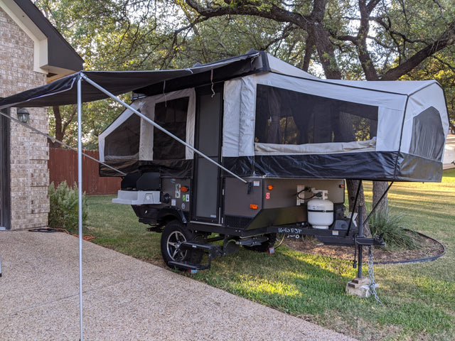 A pop-up camper, fully extended with an awning, parked on grass beside a brick house. Perfect for your next RV rental camping adventure.