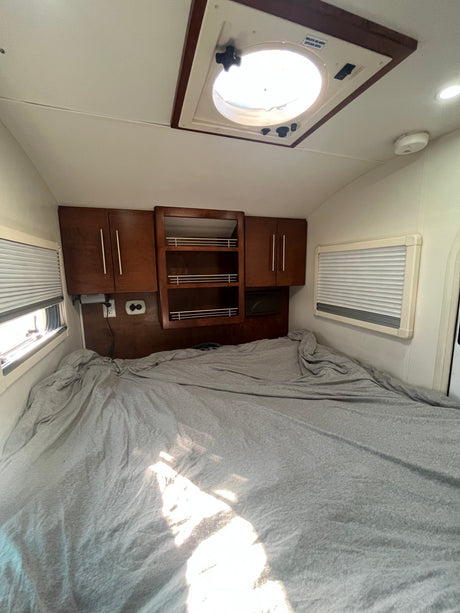 RV rental bedroom with a large bed, wooden overhead cabinets, windows with blinds, and a bright roof vent.