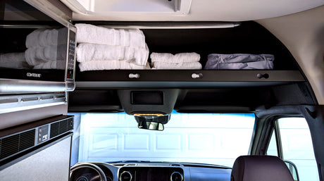 Inside an RV rental: a microwave, organized overhead storage with folded white towels, and a front view of the dashboard. Compact living.