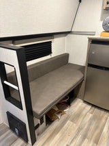RV rental interior: cozy dinette with grey faux leather bench, window blind, and compact fridge. Light wood-look floor.