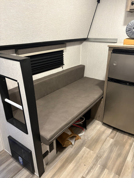 RV rental interior: cozy dinette with grey faux leather bench, window blind, and compact fridge. Light wood-look floor.