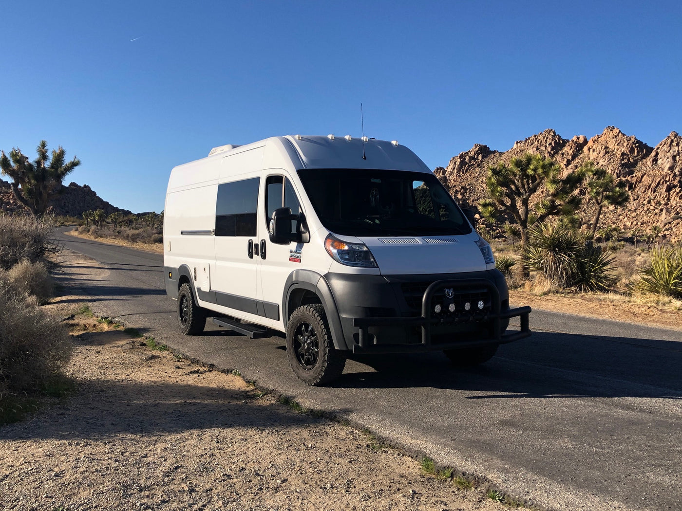 An off-road capable white RV rental van on a desert road with Joshua trees and rocky mountains under a clear sky. — photo 9