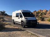An off-road capable white RV rental van on a desert road with Joshua trees and rocky mountains under a clear sky.