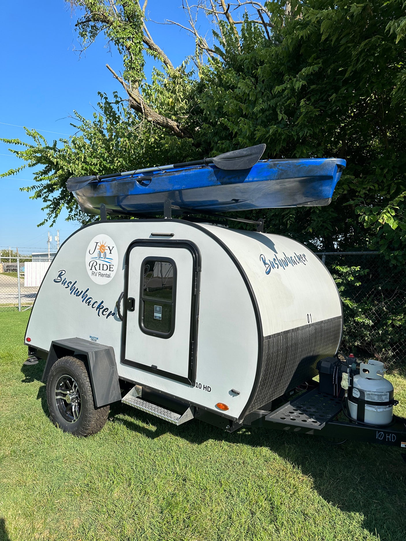 This Joy Ride RV rental Bushwhacker 10 HD teardrop camper features a blue kayak mounted on top, parked on grass.