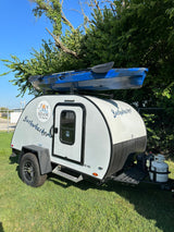 This Joy Ride RV rental Bushwhacker 10 HD teardrop camper features a blue kayak mounted on top, parked on grass.