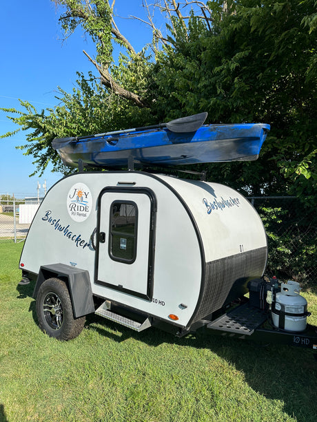 This Joy Ride RV rental Bushwhacker 10 HD teardrop camper features a blue kayak mounted on top, parked on grass.