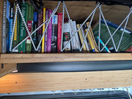 RV rental bookshelf filled with nature, hiking, and travel field guides, secured by bungee cords.