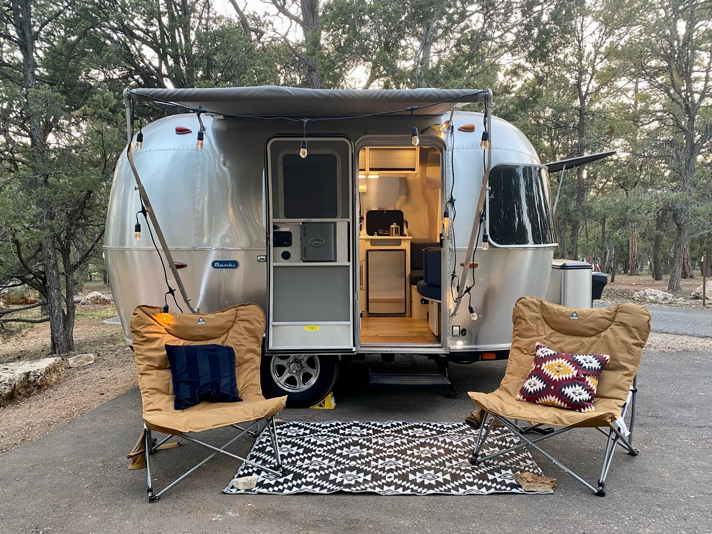 A silver Airstream Bambi rv rental is parked with an awning and string lights. Two chairs and a mat create a cozy outdoor setup.