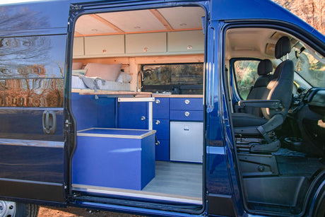 This blue camper van conversion shows a complete interior with bed, kitchen, storage, and front seats, ready for RV rental.