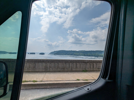 Capturing a wide river with islands & forested hills, seen through an RV rental window from a bridge under a cloudy sky.