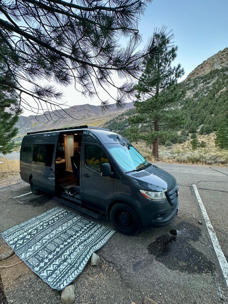 Discover nature with an RV rental! This campervan is parked by pine trees in mountains, with an outdoor rug out. — photo 18