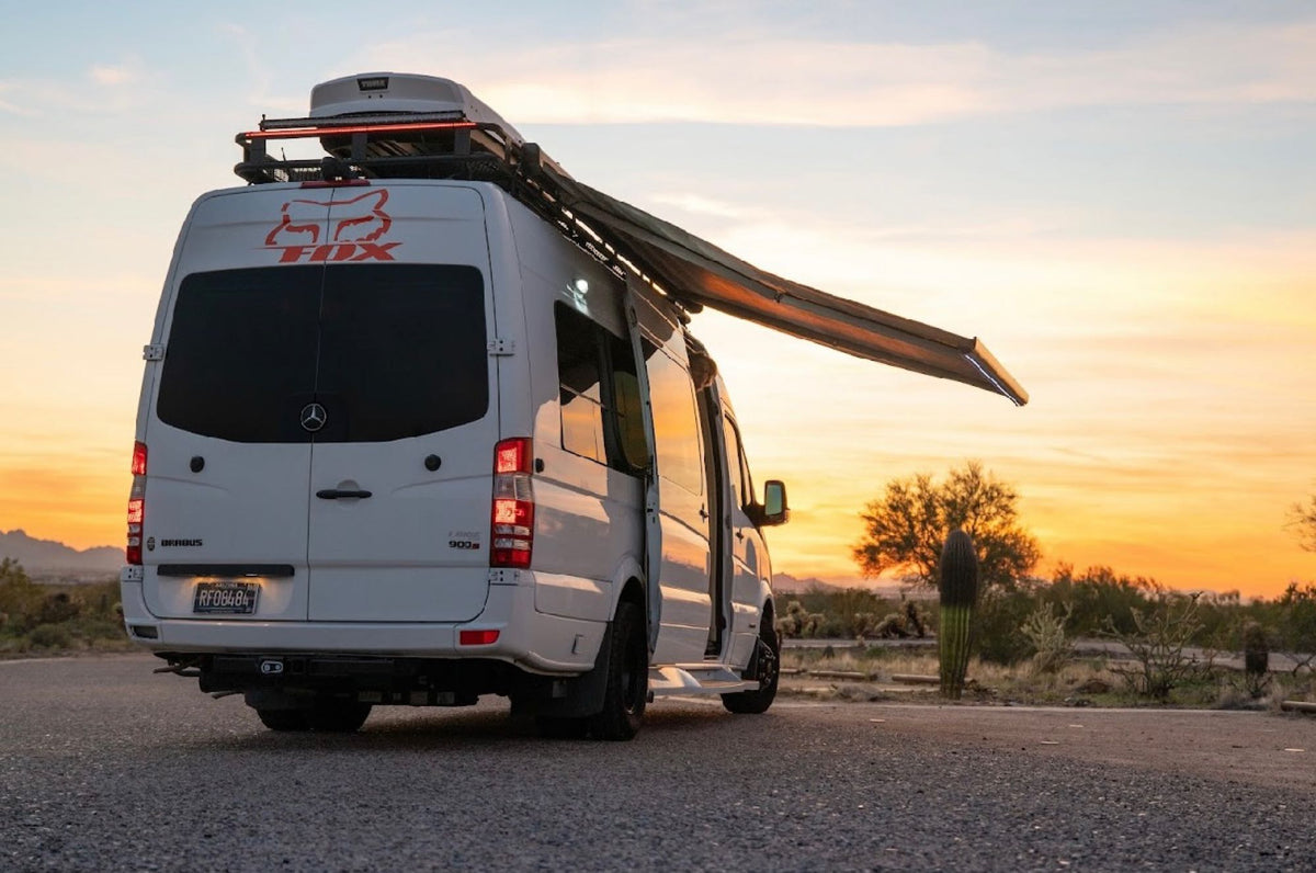 Experience desert adventures in this white custom Sprinter RV rental. Its extended awning and roof storage are ready for travel.|White camper van