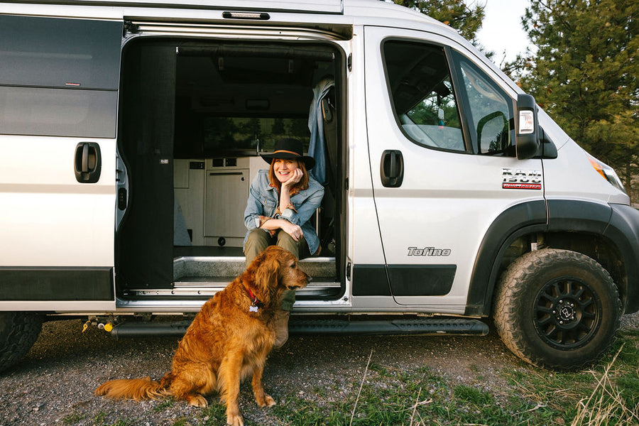 Smiling woman and her dog enjoying an RV rental adventure in their Tofino camper van, parked with the side door open.