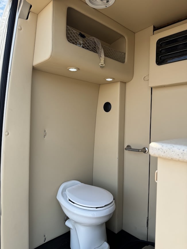 RV rental interior: Compact bathroom area featuring a white toilet, beige padded walls, overhead lights, and mesh storage. — photo 17