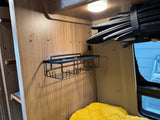RV rental interior: wood paneling, wall-mounted wire shelf for storage, overhead light, rolled shade, and yellow pillow.