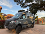 A sleek grey Mercedes Sprinter campervan, a Winnebago Revel 4x4, with off-road tires under a pine tree. Ready for your rv rental adventure!