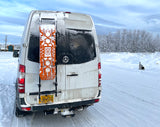 Alaska RV rental van in winter, covered in snow with traction boards, parked in a snowy landscape with a dog nearby.