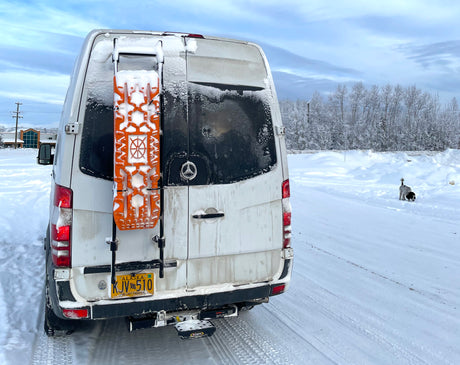 Alaska RV rental van in winter, covered in snow with traction boards, parked in a snowy landscape with a dog nearby.