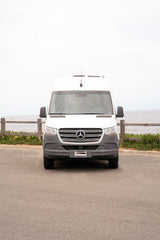 Front view of a white Mercedes-Benz Sprinter camper van, an RV rental, parked on asphalt by the ocean.