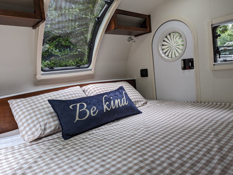 Cozy RV rental interior featuring a comfortable bed with checkered bedding, a "Be kind" pillow, and a clear window view of nature.