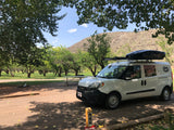 White RV rental van with rooftop cargo box parked at shaded campsite 68, surrounded by an orchard.
