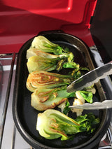 Halved bok choy is sautéing in a black pan with tongs, possibly on an RV rental outdoor stove.