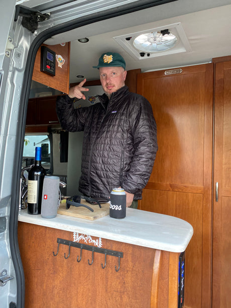 Inside an RV rental: a man making a peace sign beside a kitchen counter with wine, speaker, and a casual drink.