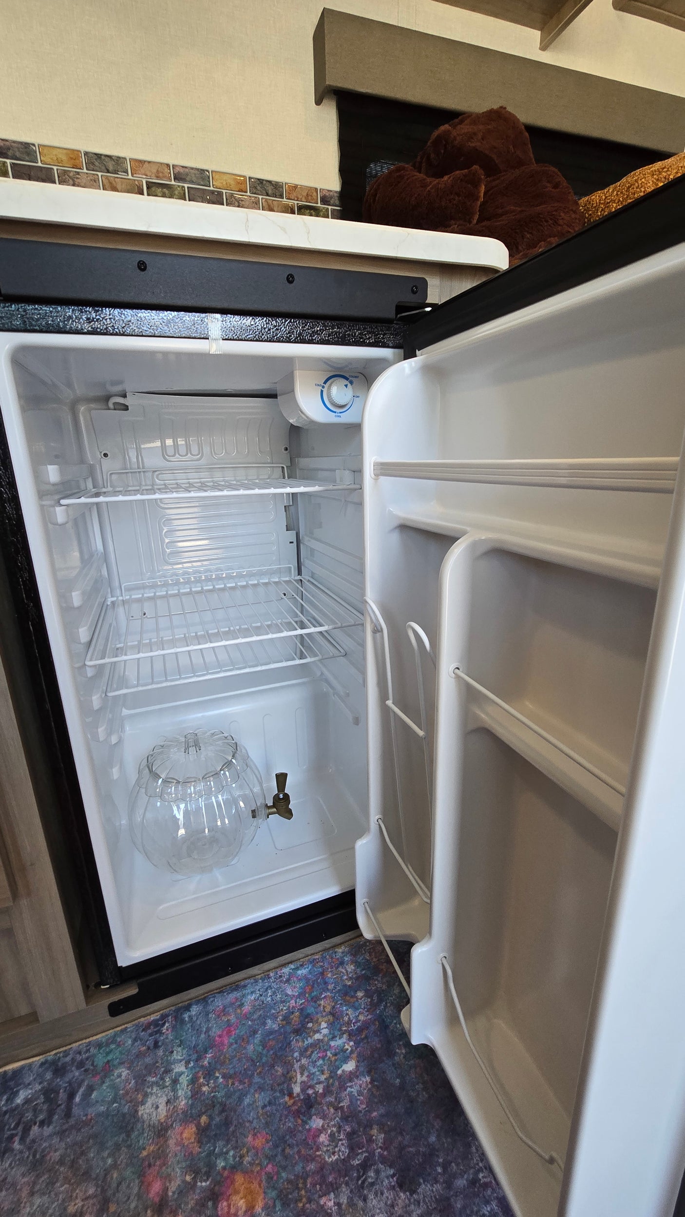 Empty mini-fridge in an rv rental, with wire shelves and a clear, pumpkin-shaped beverage dispenser.