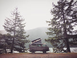 A dark camper van with pop-top, perfect for rv rental, parked on dirt by a misty lake with tall trees & forested mountains.