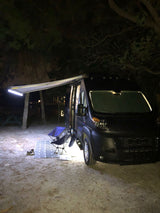 Dark camper van rv rental at night, with an illuminated awning, chairs, and mat set up for a cozy outdoor evening camp.
