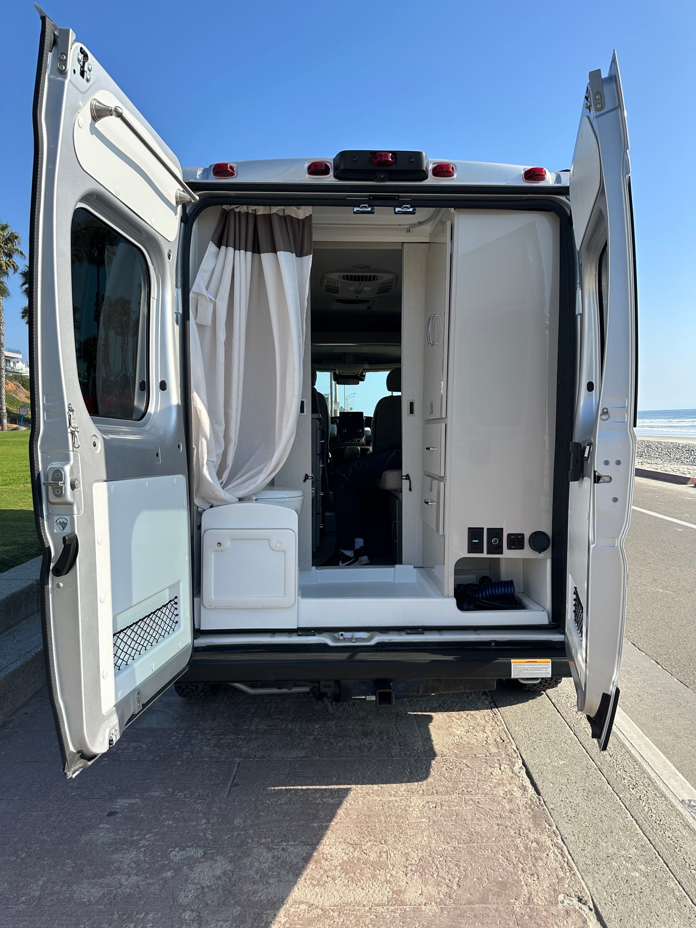 This rv rental camper van's open rear doors reveal its versatile interior with a stunning beach view.