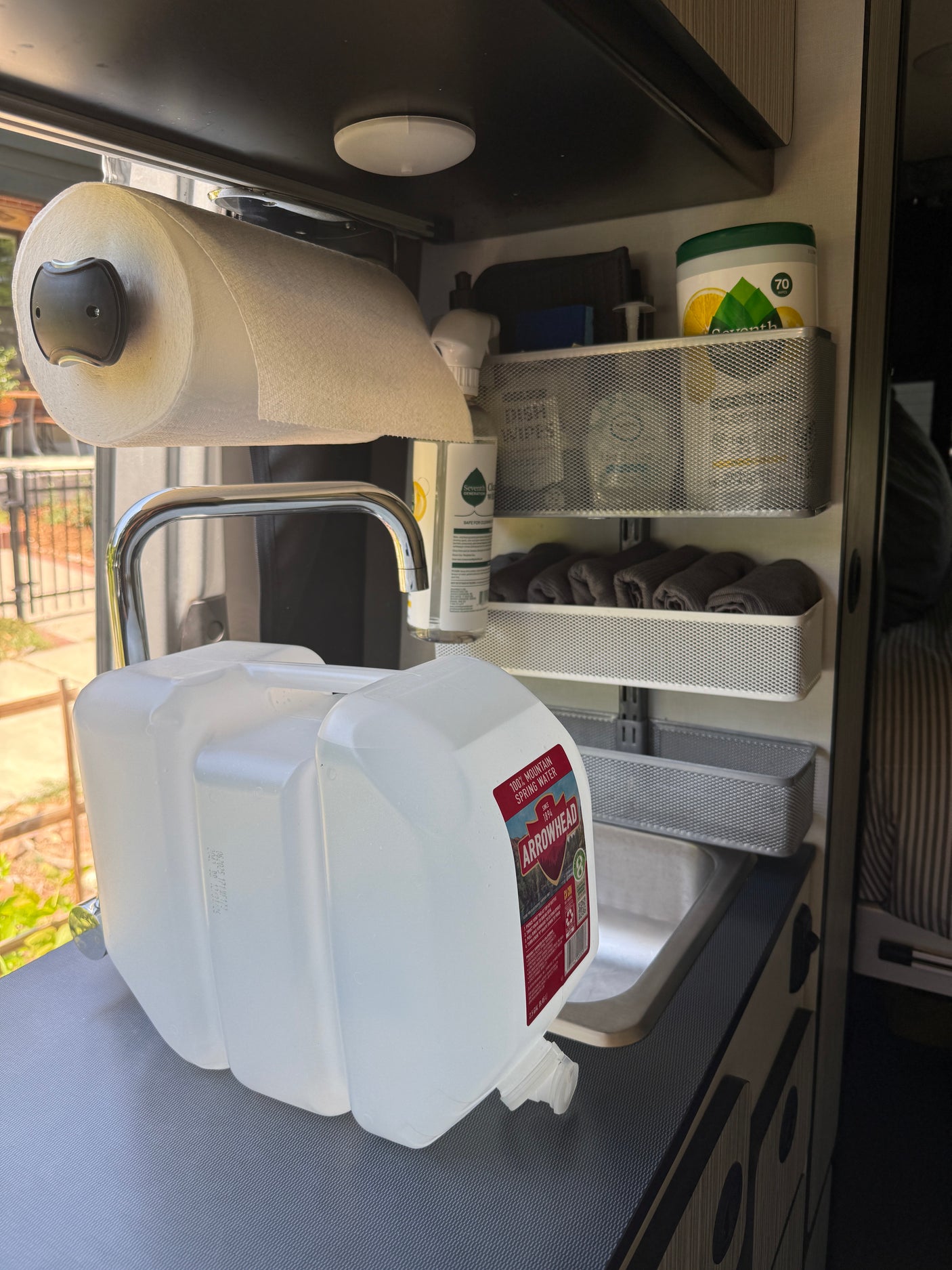 Efficient RV rental kitchen setup: water jugs by the sink, paper towels above, and shelves with cleaning supplies and rolled towels.