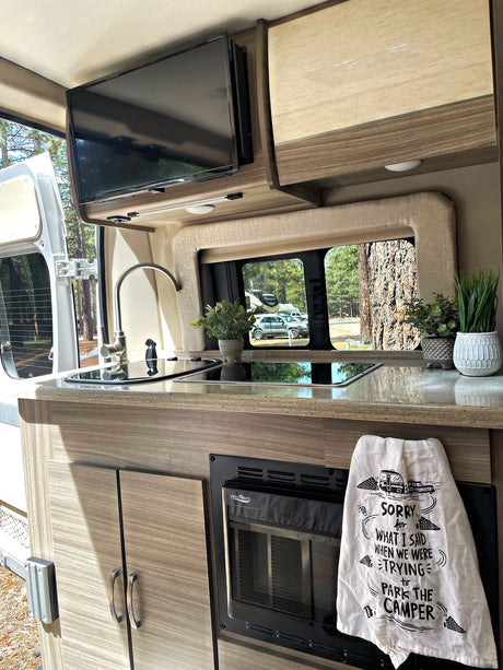 Inside a modern rv rental kitchen: a sleek counter with sink & stovetop, microwave below, TV above, and a playful towel.