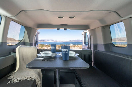 Inside an rv rental: dining table for two with Escape Campervans cups, offering a stunning lake and mountain view.