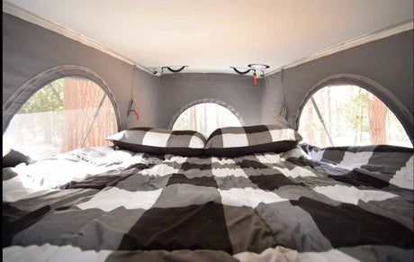 Cozy RV rental bed with buffalo plaid bedding, offering forest views through arched windows. Ideal for glamping trips.