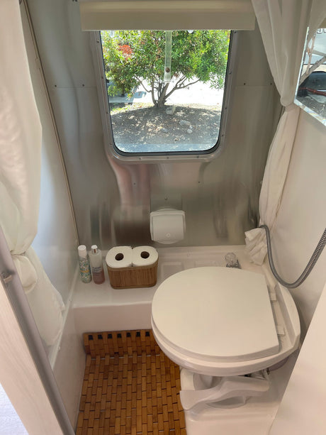 Shiny RV rental wet bath: toilet, shower, slatted floor, window view to trees.