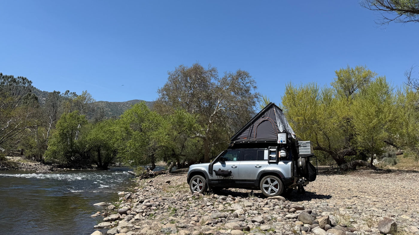 Silver Land Rover Defender with rooftop tent, set up like an RV rental by a rocky river. Perfect for adventure trips! — photo 6