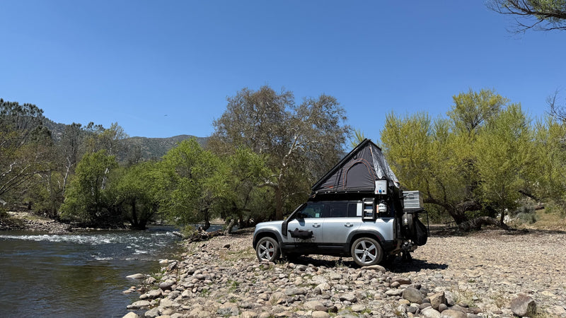 Silver Land Rover Defender with rooftop tent, set up like an RV rental by a rocky river. Perfect for adventure trips! — photo 6