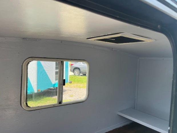 Inside a light gray RV rental interior with a window, overhead vent, and practical storage shelf.