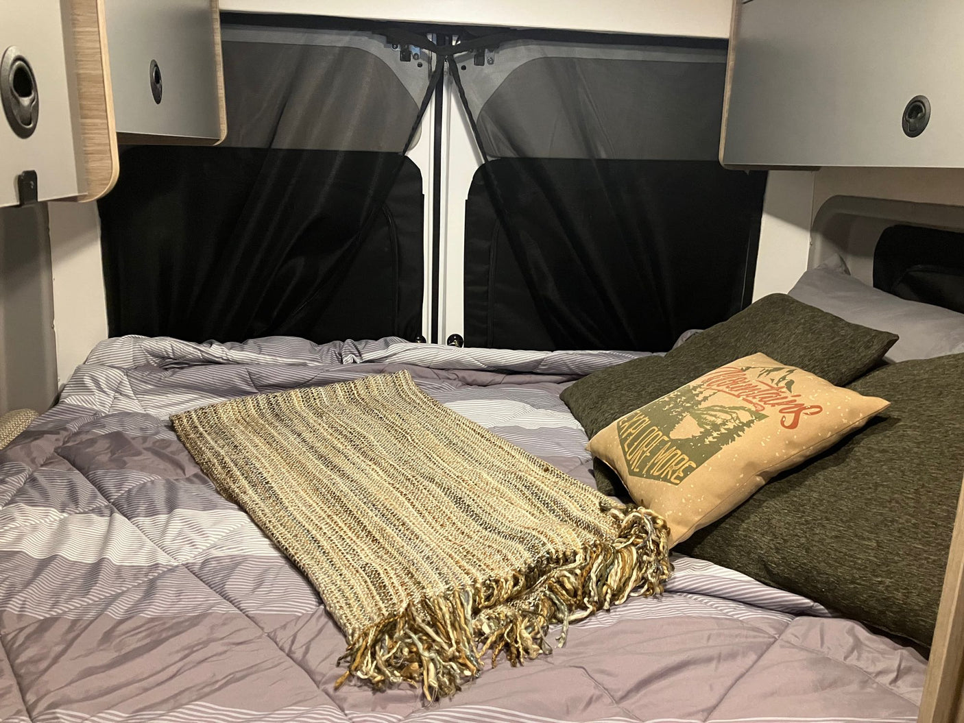 Inside an RV rental: a soft bed with a patterned comforter, fringe blanket, and comfy pillows, framed by overhead cabinets & privacy screens.