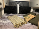 Inside an RV rental: a soft bed with a patterned comforter, fringe blanket, and comfy pillows, framed by overhead cabinets & privacy screens.