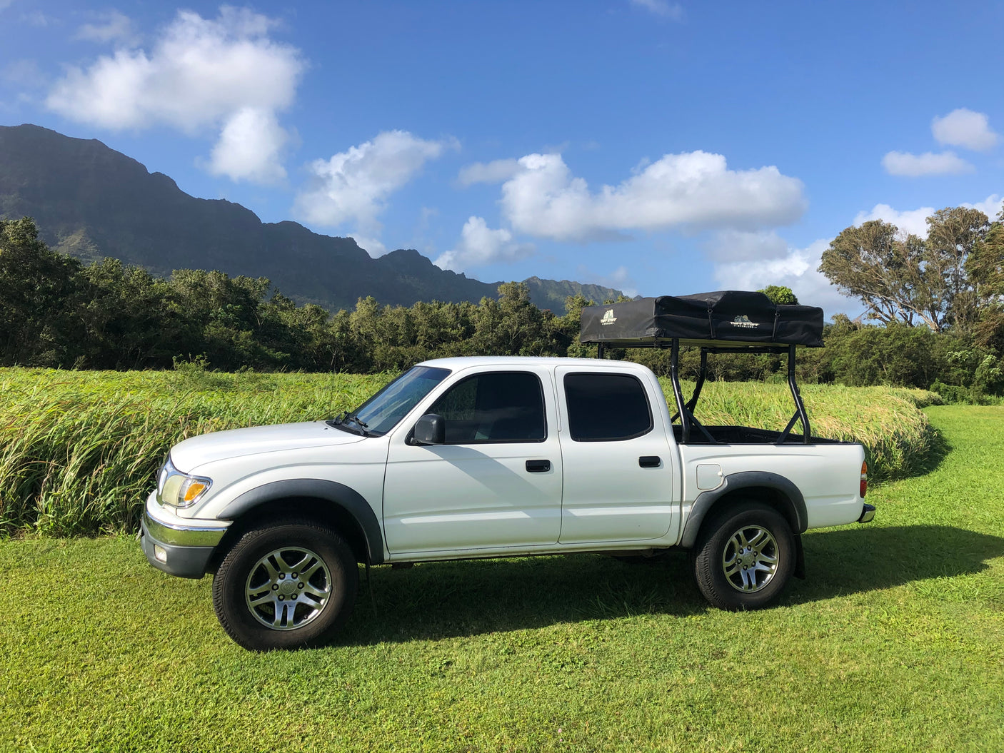 White pickup truck with a rooftop tent for rv rental, parked in a lush field with mountains in the background.