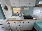 Kitchen and sleeping area inside an RV rental, showing a compact sink, counter, cabinets, and a cozy bed.