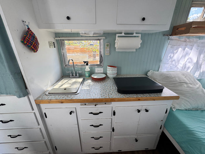 Kitchen and sleeping area inside an RV rental, showing a compact sink, counter, cabinets, and a cozy bed.